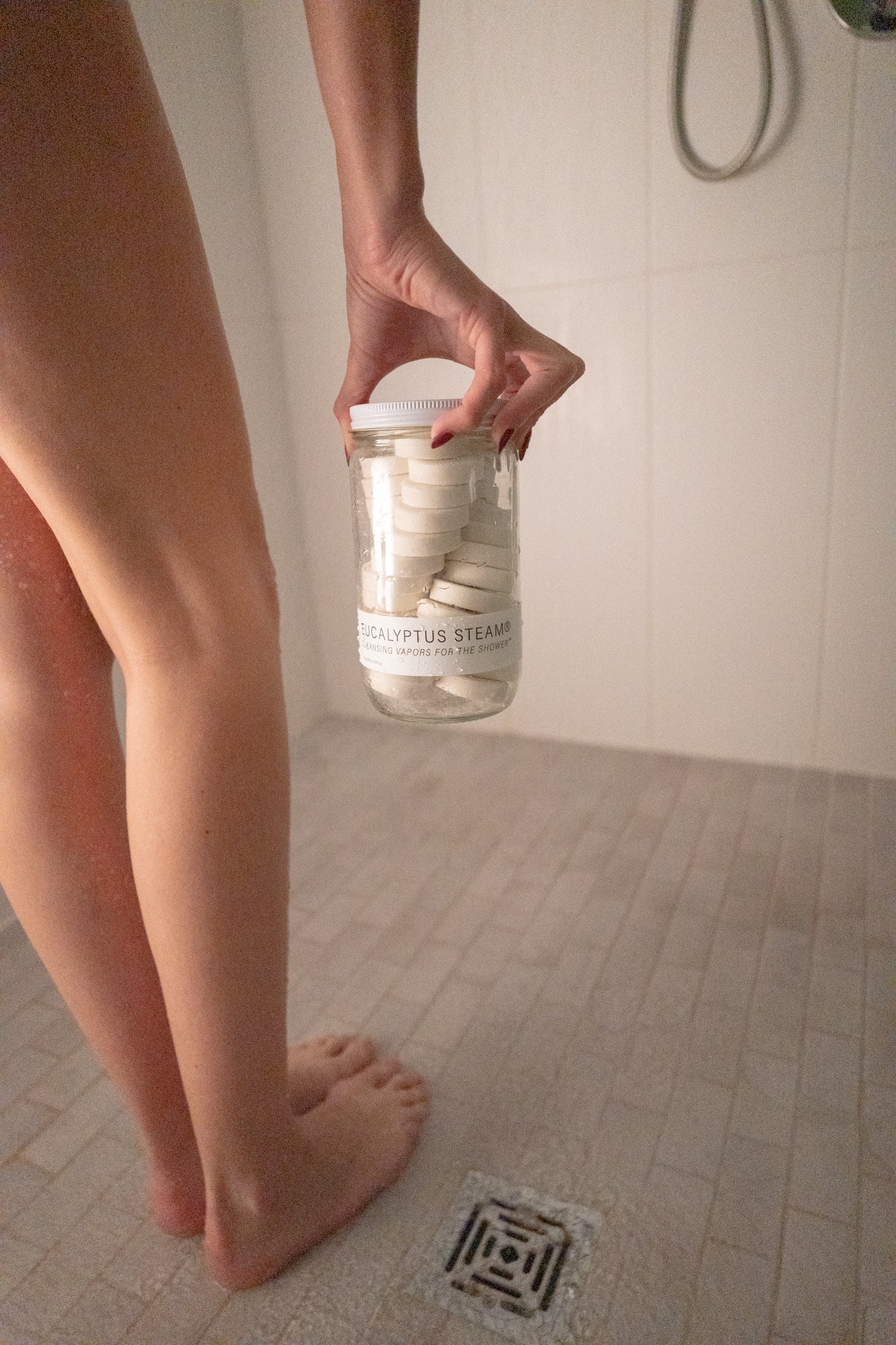 Hand holding a jar of white shower vapor tablets near a shower drain