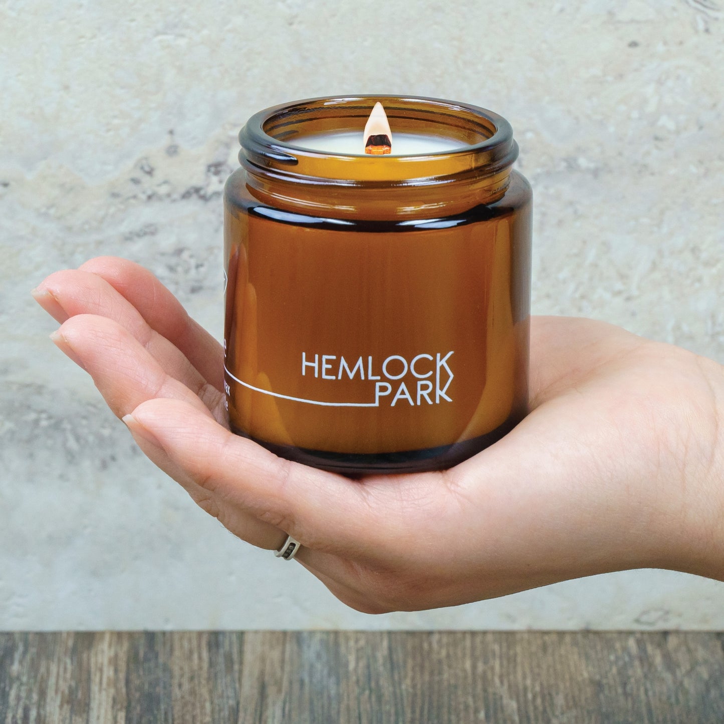Amber glass coconut wax candle jar with wick lit, held in hand, labeled Hemlock Park