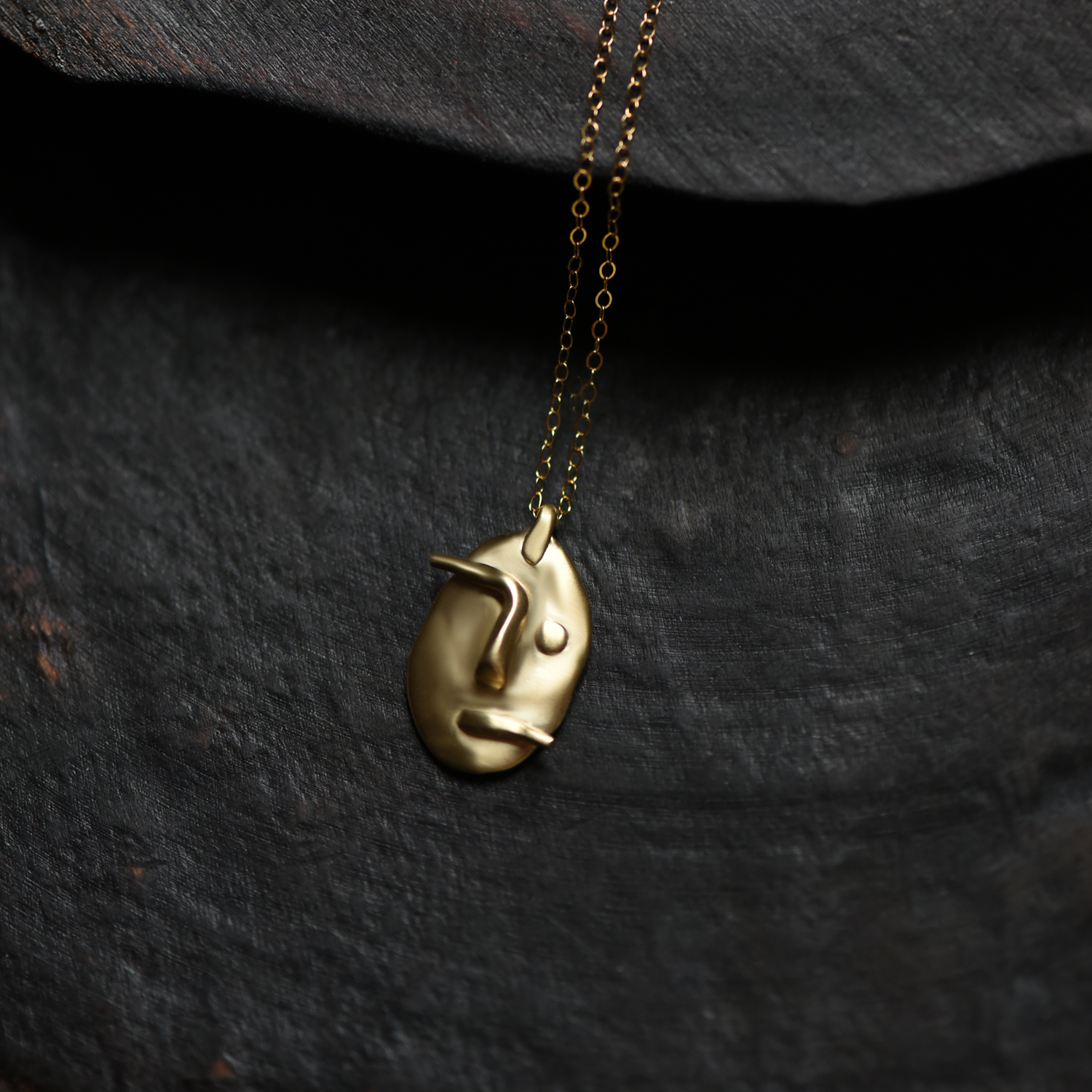 Gold-tone face pendant necklace with round pendant and dangling chain on dark cloth