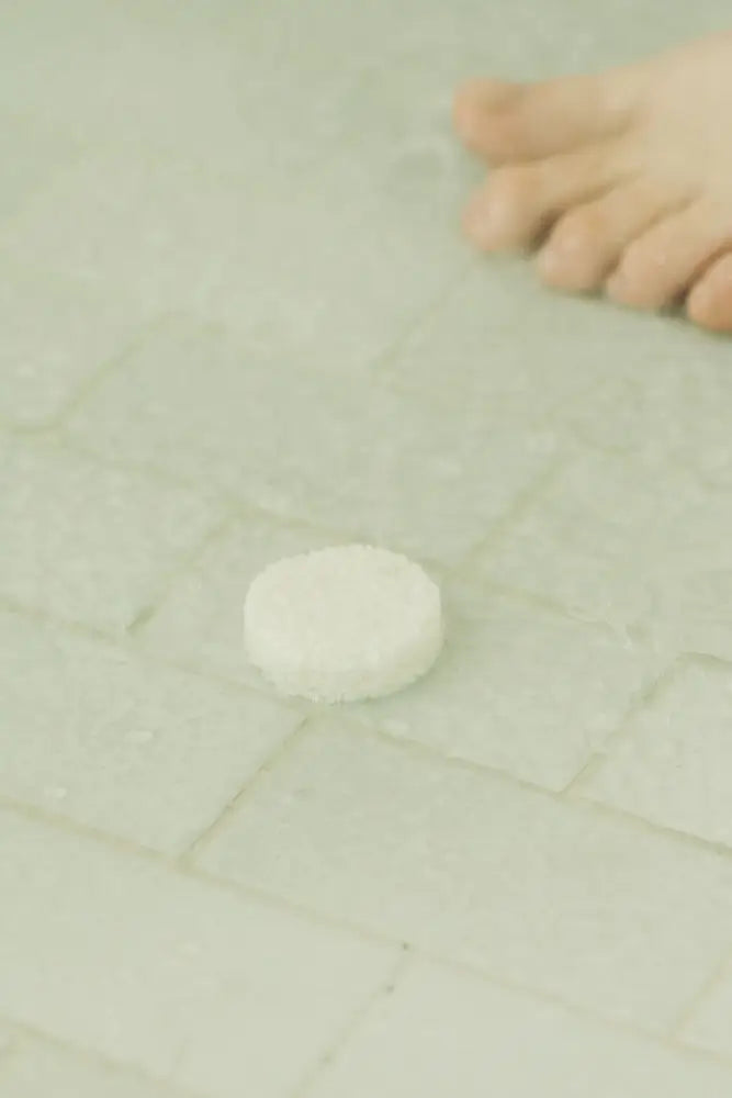 White shower steam tablet on light tile floor with hand nearby