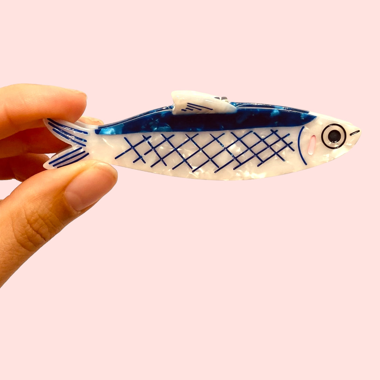 Fish-shaped hair claw clip, blue grid on cream body, held by a hand
