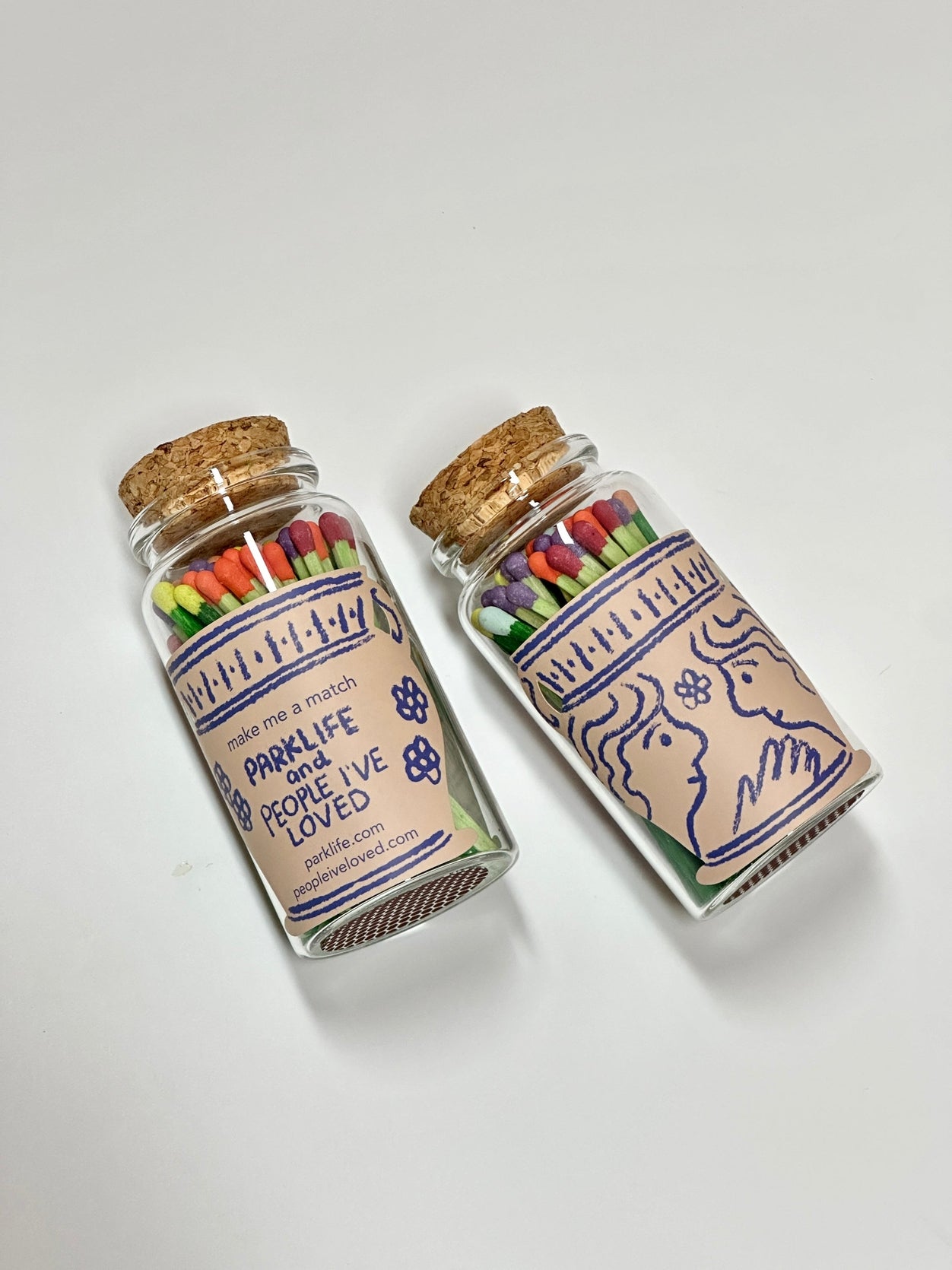 Two decorative match jars with cork lids and illustrated labels, filled with colorful matches