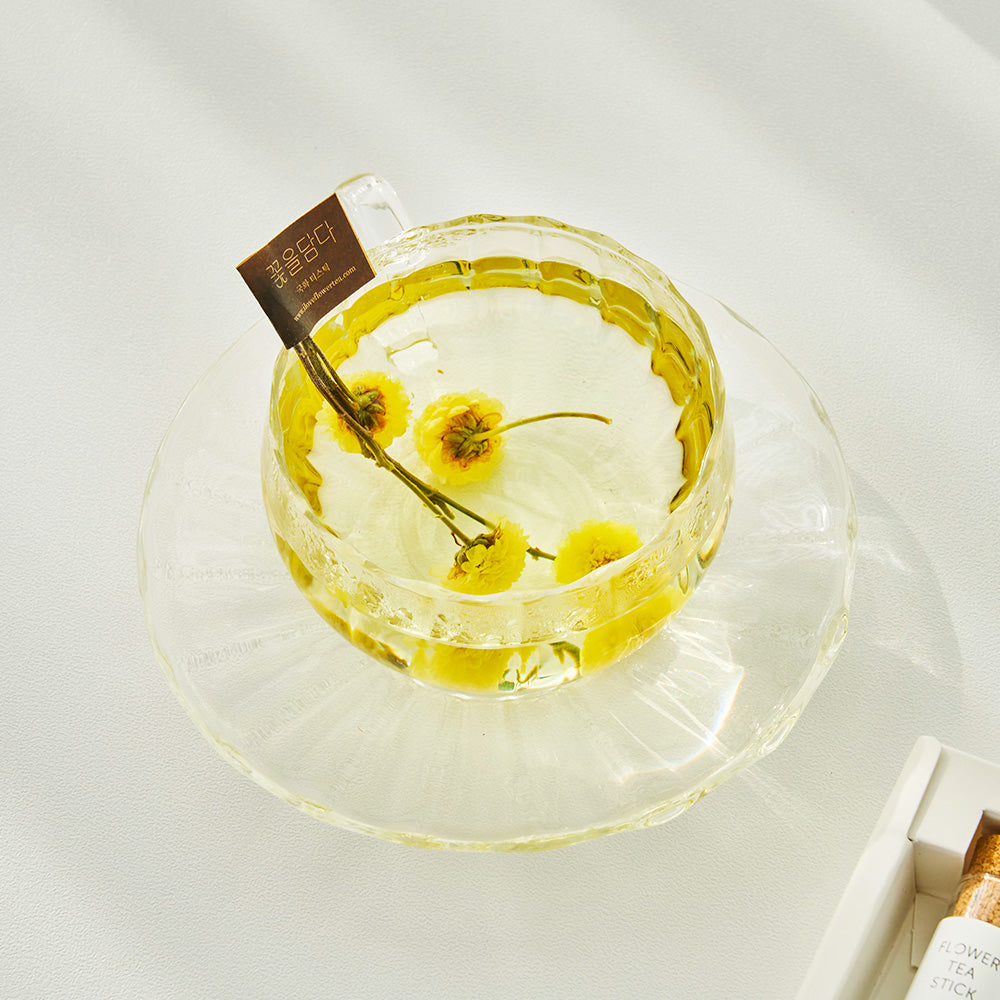 Glass bowl of golden tea with yellow dried flower petals and a tea stick sachet