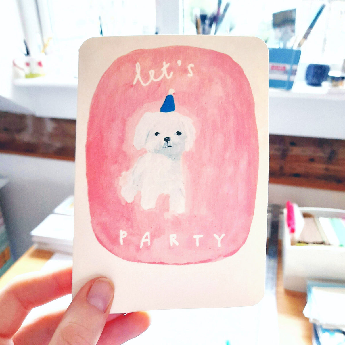Dog party greeting card with white dog in blue hat inside pink watercolor circle and “PARTY” text