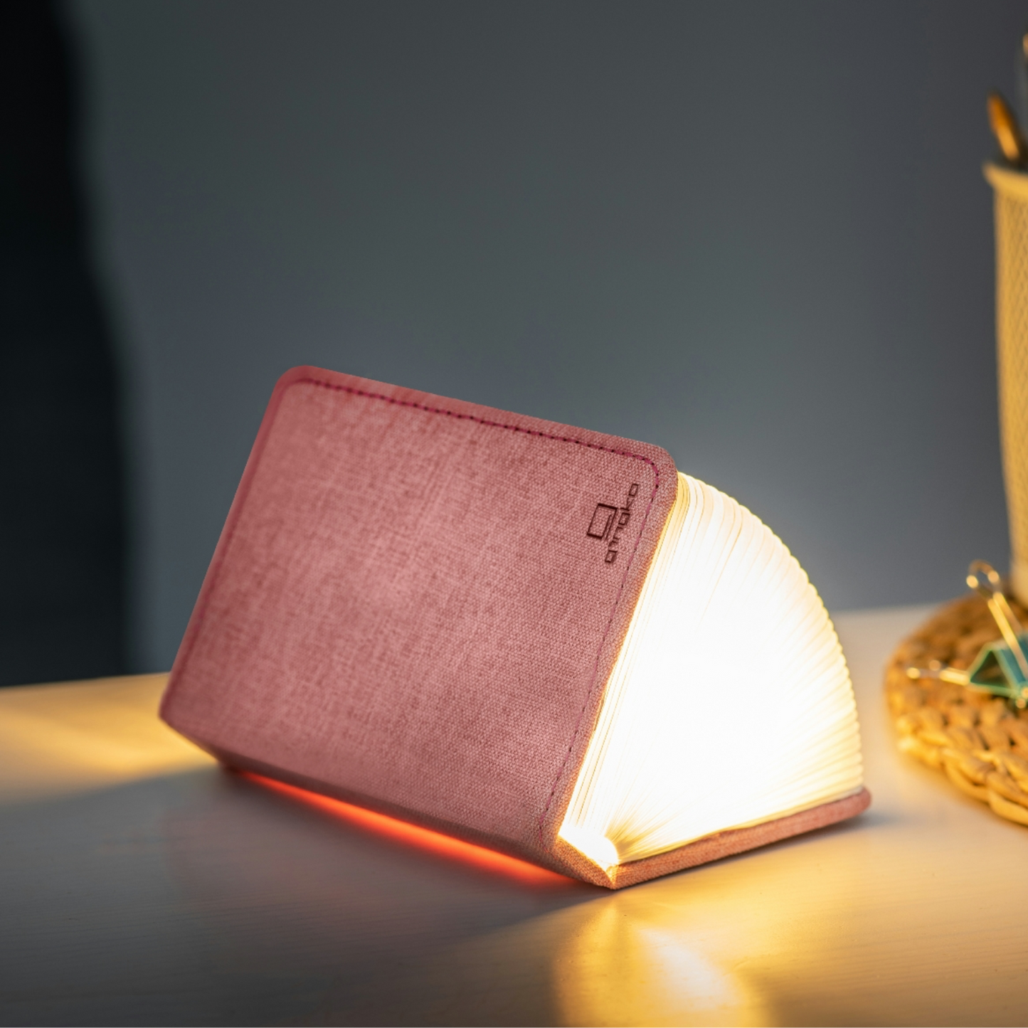 Blush pink linen smart booklight with warm glowing pages on an angled base