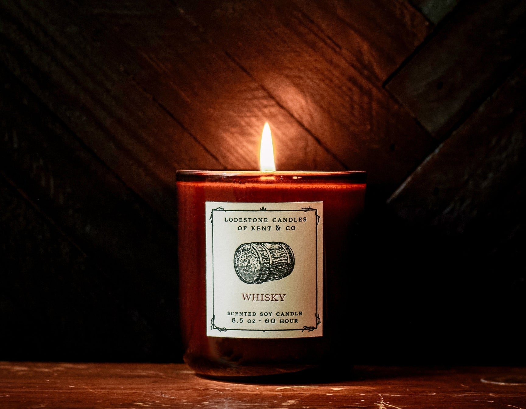 Amber glass jar soy candle with lit wick and cream label on dark wooden surface