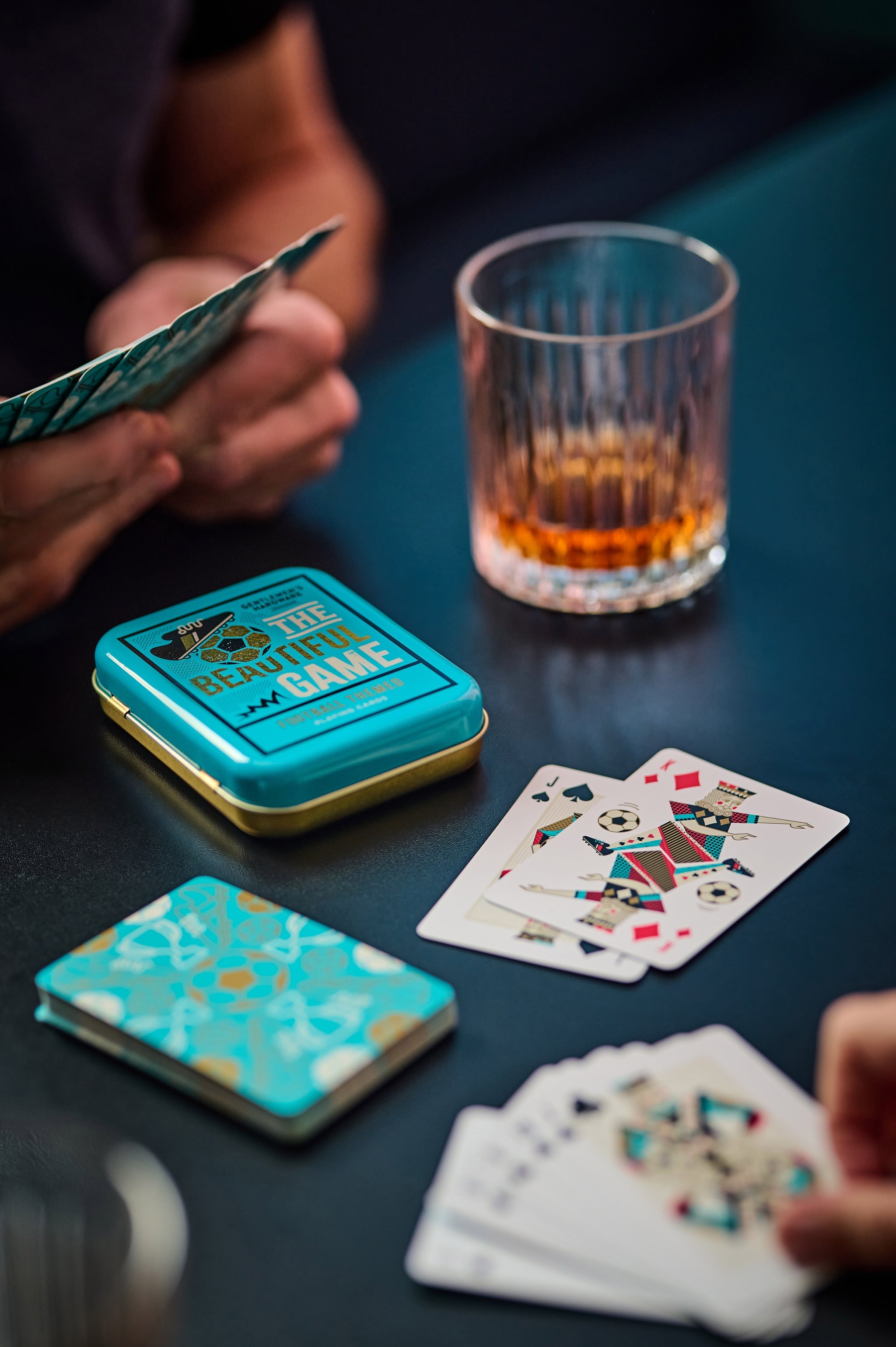 Football-themed playing cards with teal tin and face-up cards, played by hands on table.