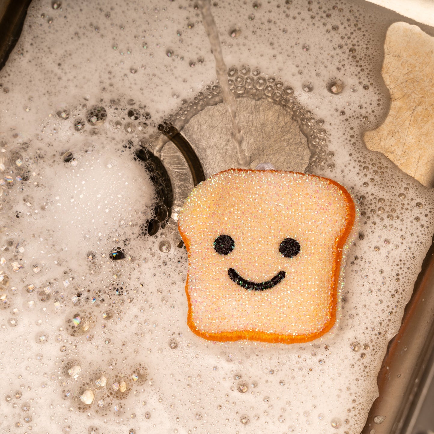 Orange toast-shaped scrub sponge with smile face, in sink with bubbles