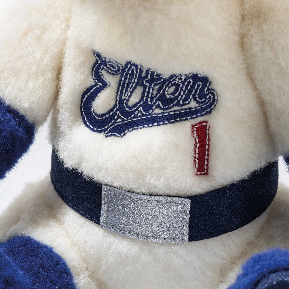 Teddy bear chest detail: stitched “Elton” patch on cream shirt with blue belt