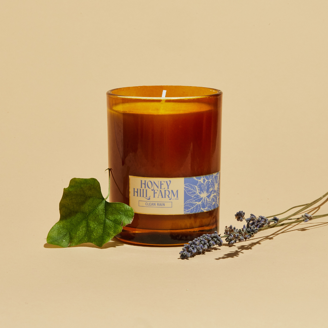 Amber glass candle with 'Honey Hill Farm' label, with leafy greens and lavender sprigs