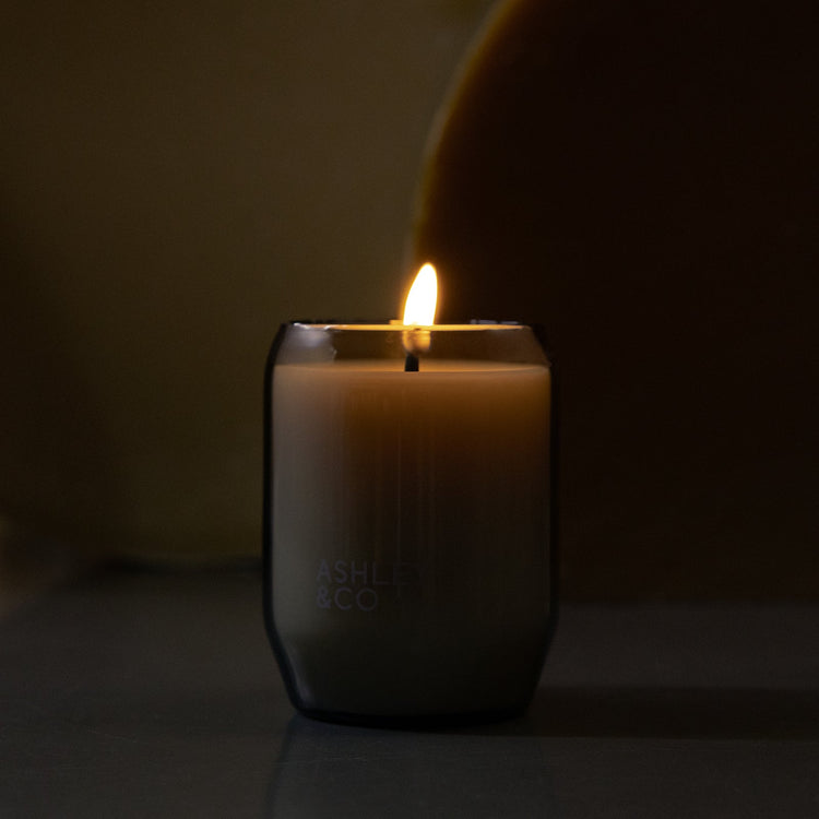 Waxed perfume candle with lit flame inside smoky olive glass jar
