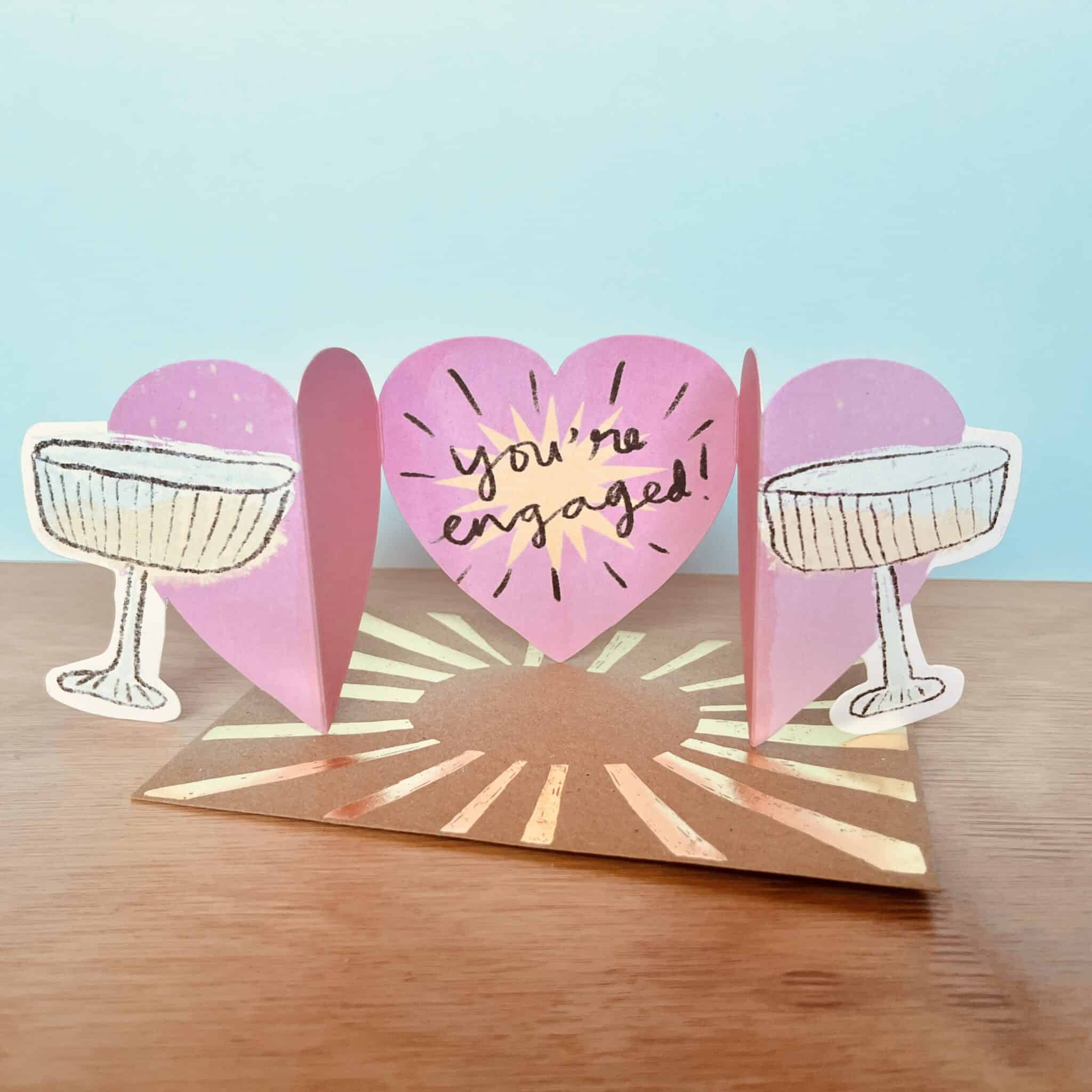 Clinking glasses engagement greeting card with pink heart and "You're engaged!" text