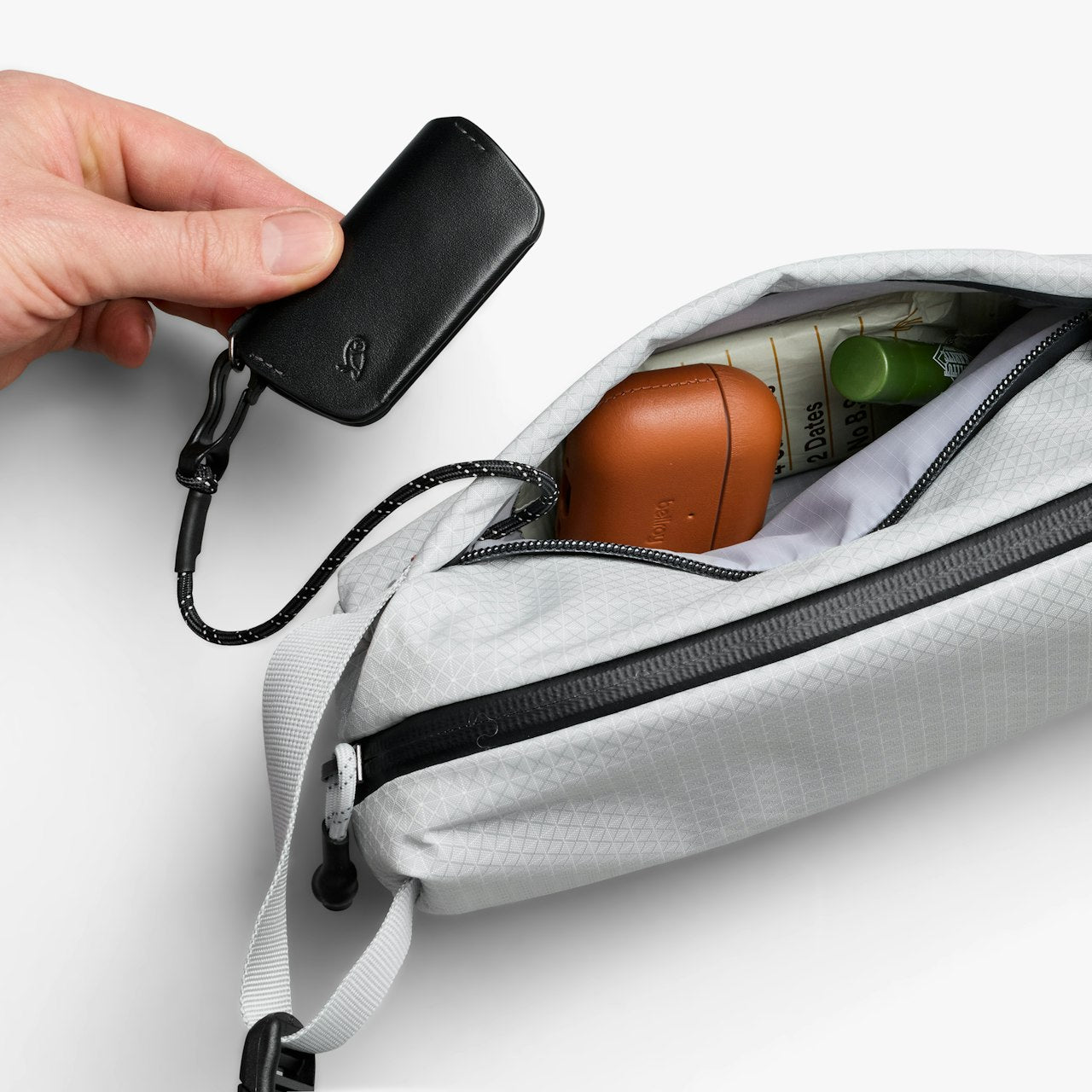 Light gray mini sling bag with zip open and black keychain pouch held by hand.