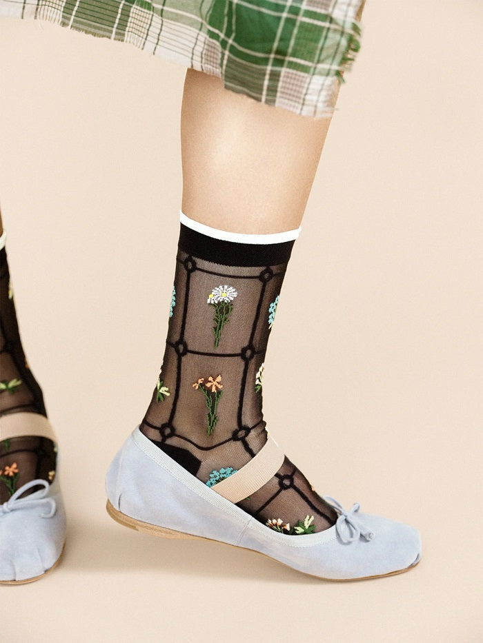 Black sheer crew socks with floral vine and daisy motifs, worn on white flats