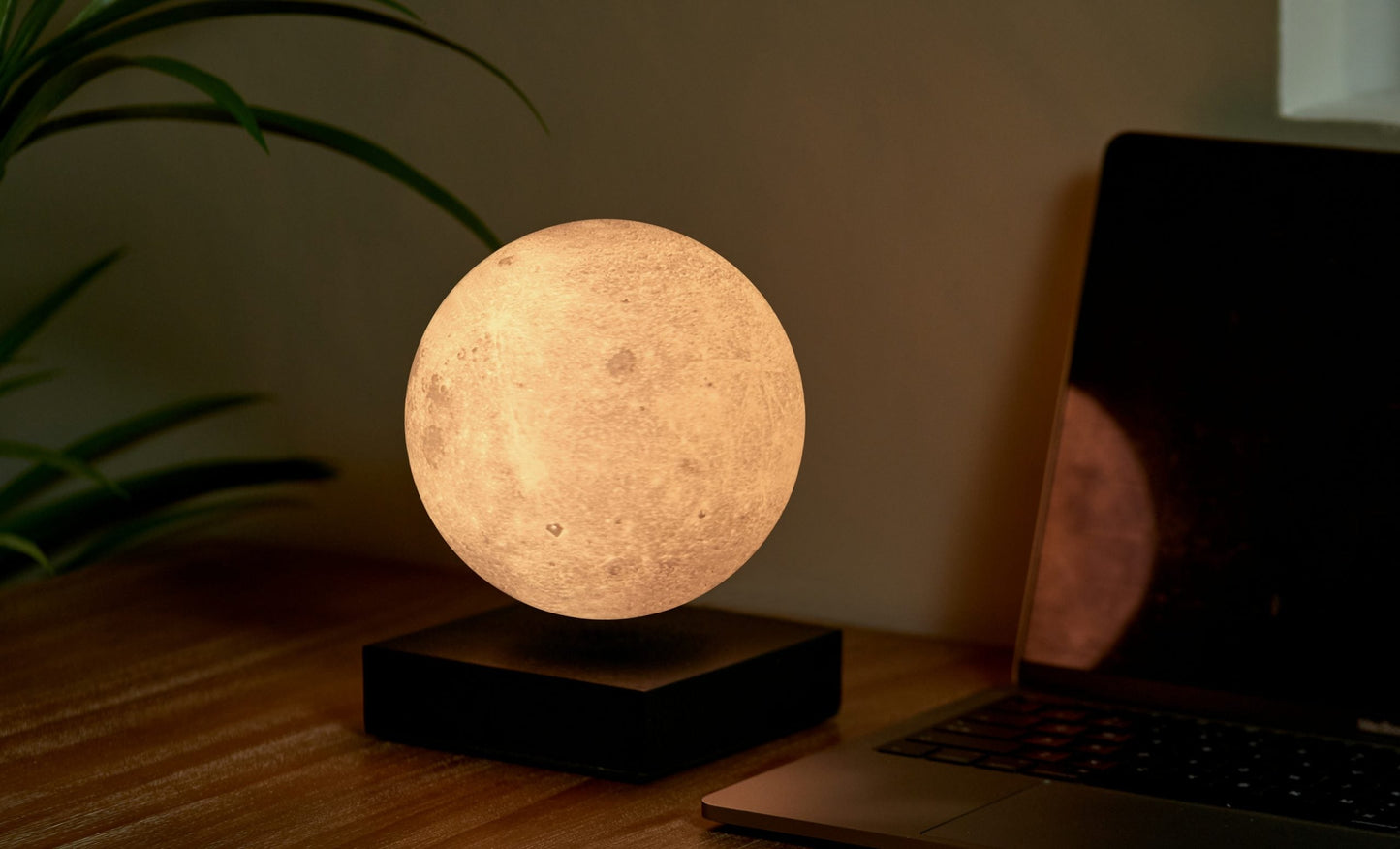 Moon lamp with warm glowing sphere on black base beside laptop