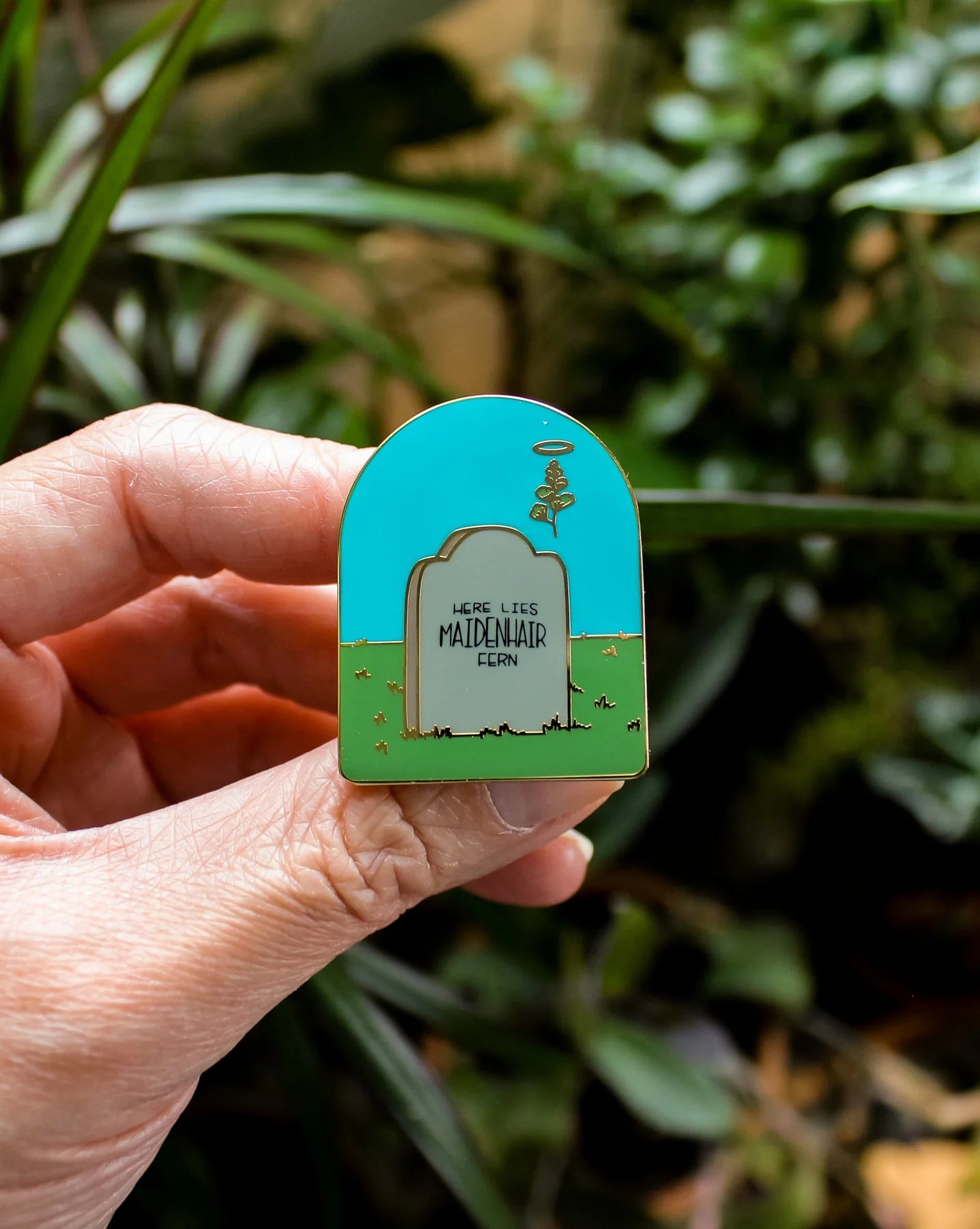 Maidenhair fern grave enamel pin in hand, turquoise arch with gravestone and fern tree motif