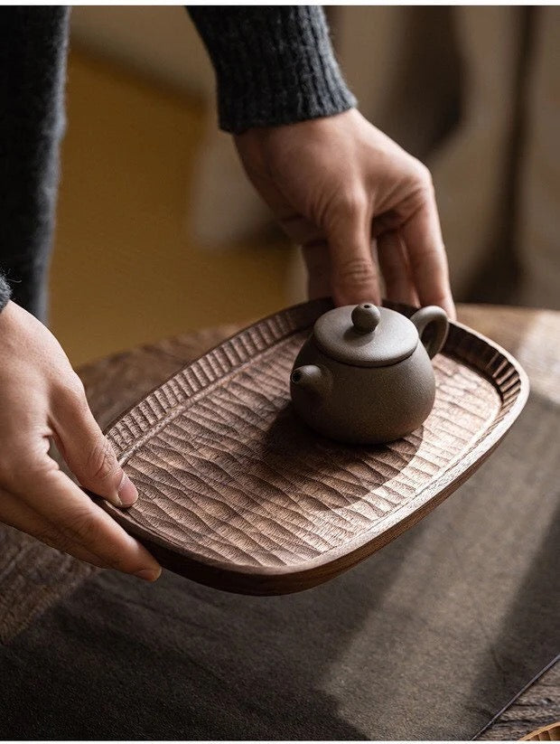 Wooden tea tray with textured center and carved rim, with teapot on top while being placed