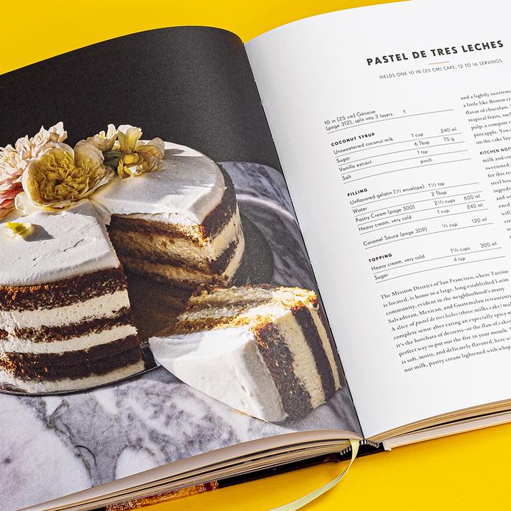 Cookbook open to PASTEL DE TRES LECHES, with cake slice photo and recipe list
