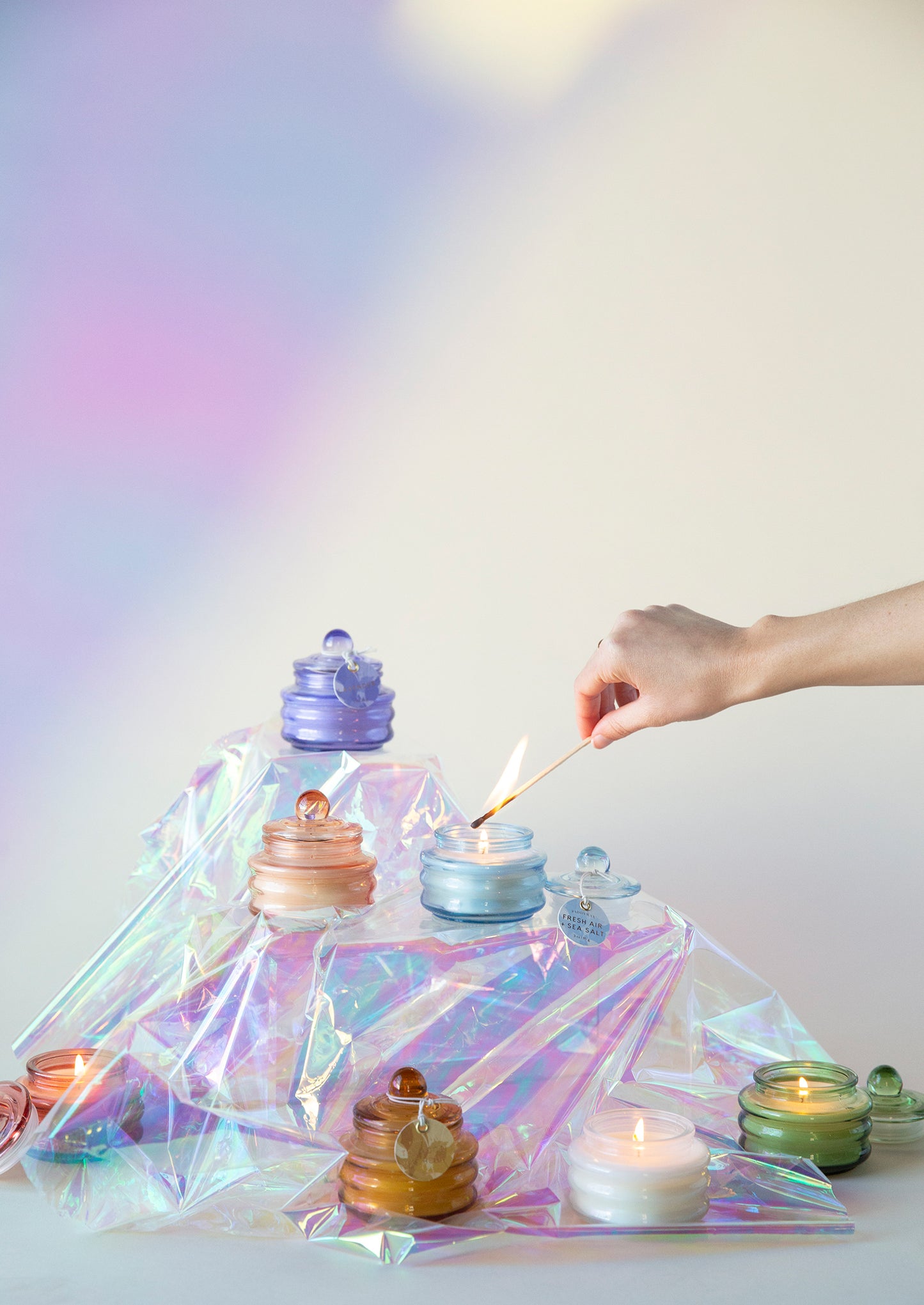 Beam candle jars with burning wicks on an iridescent tray; hand lights one